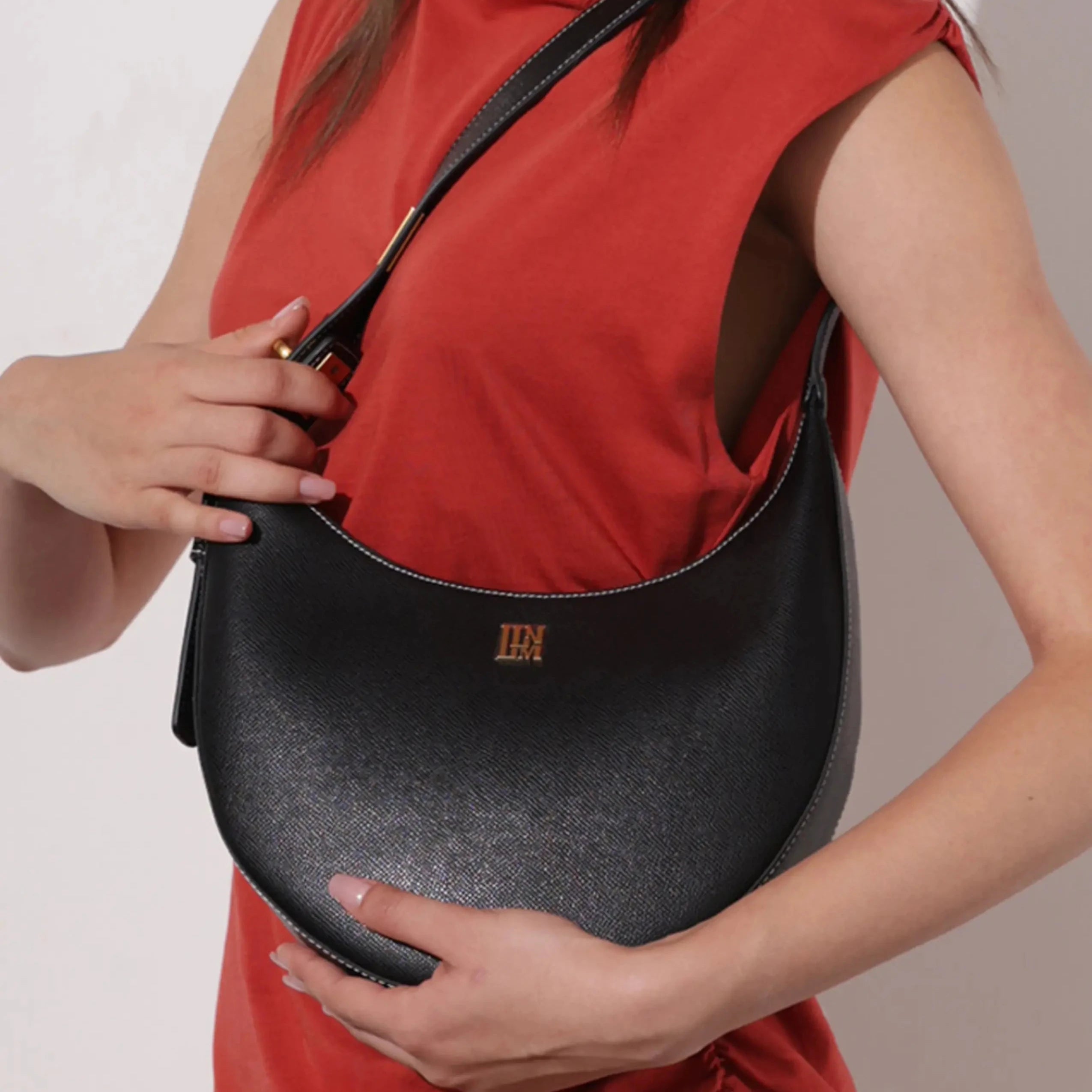 Black leather crescent shoulder bag with gold logo worn by woman in red top, minimalist luxury handbag for modern everyday fashion styling