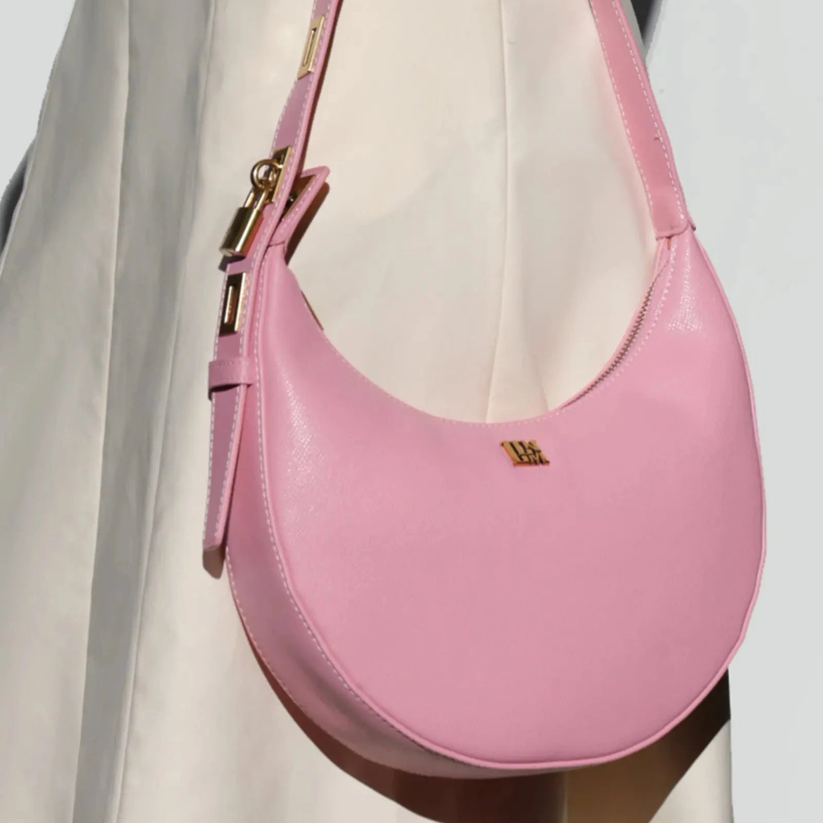 Pink leather crescent shoulder bag with gold hardware held by woman, minimalist half moon purse in soft pastel color for elegant everyday styling