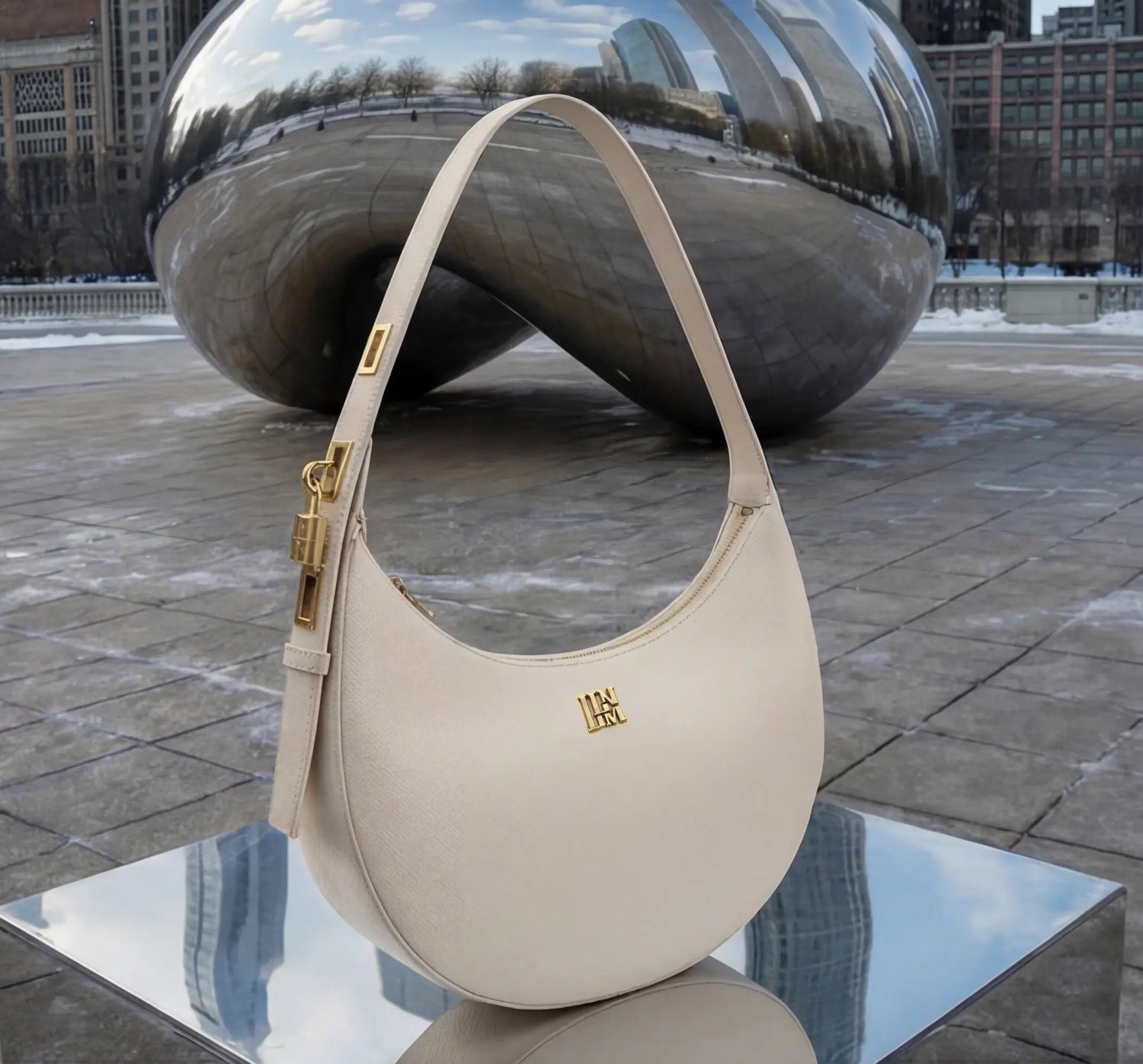 White crescent leather shoulder bag with gold hardware displayed on reflective surface near urban sculpture, luxury minimalist handbag product detail shot.