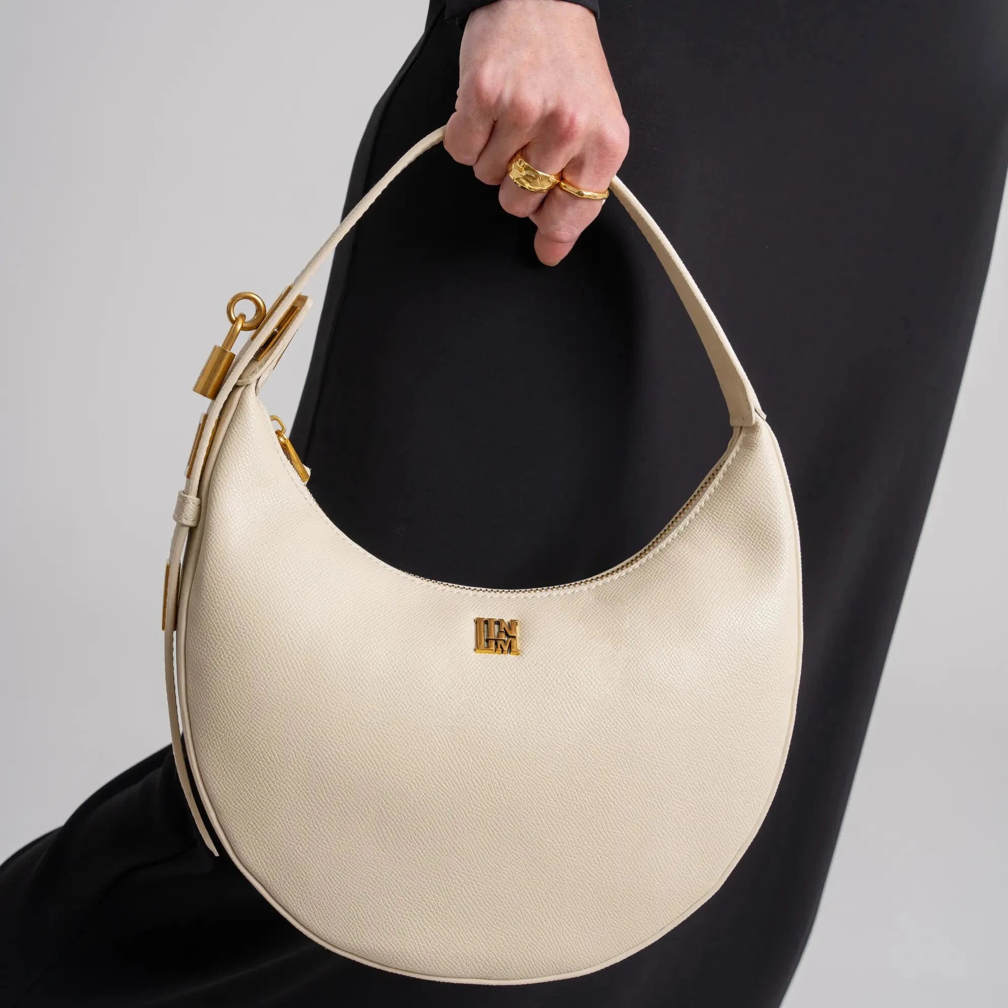 White leather crescent shoulder bag with gold hardware held by woman in black outfit, minimalist half moon handbag with clean architectural design