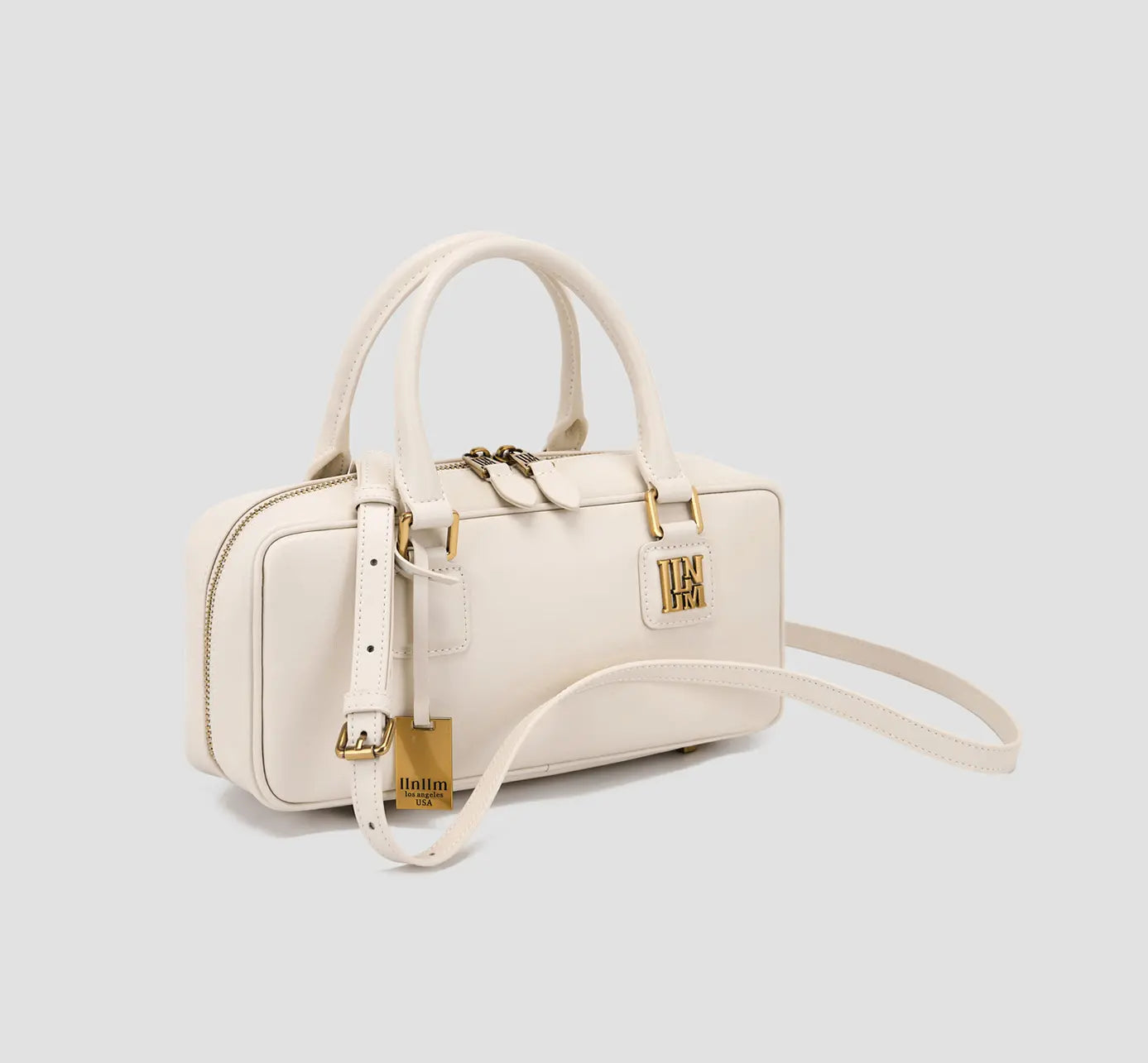 The angled view of a cream structured handbag with a shoulder strap and gold-tone branding accents.
