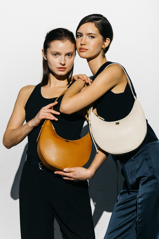 Models showcase llnllm moon-shaped shoulder bags in tan and cream leather, styled with black outfits for a sleek, modern over-the-shoulder look.