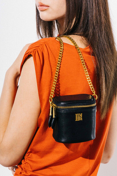 Model in orange top wears llnllm black leather mini crossbody bag with gold chain strap and zipper closure, perfect for chic hands-free styling.
