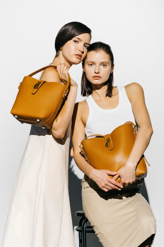 Two models showcase llnllm tan leather bucket bags with gold hardware, styled with neutral dresses for a chic designer handbag look.