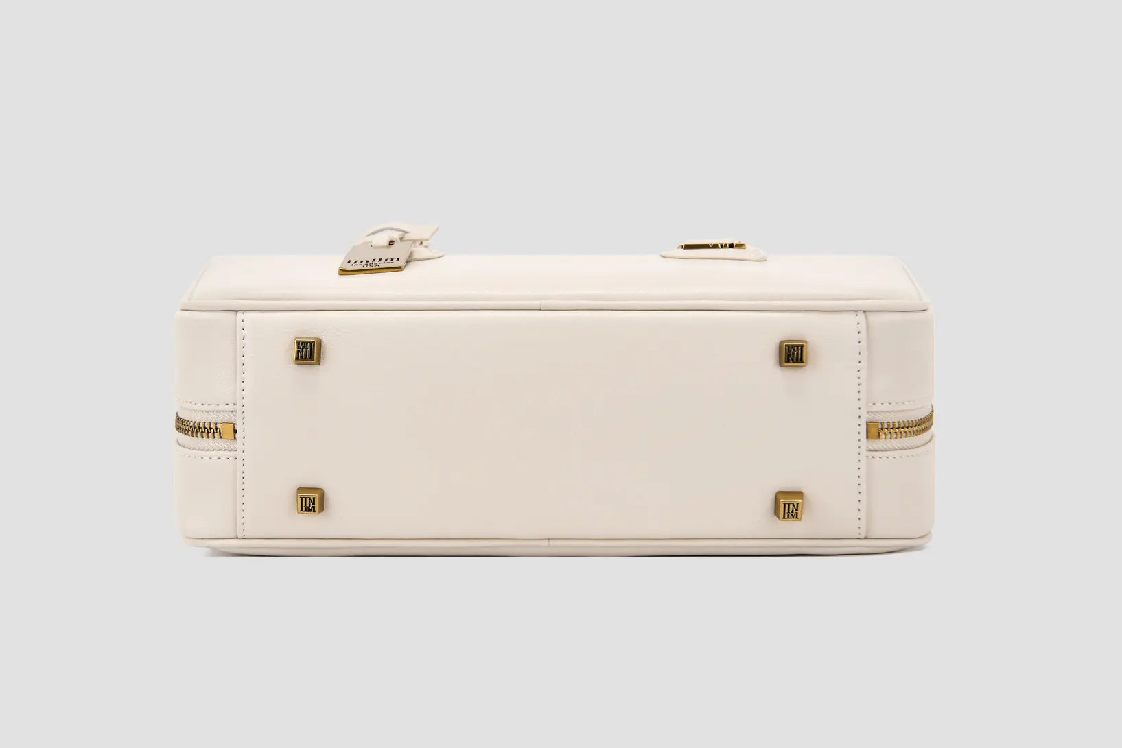 The bottom of structured white leather handbag with gold logo feet and dual side zippers.
