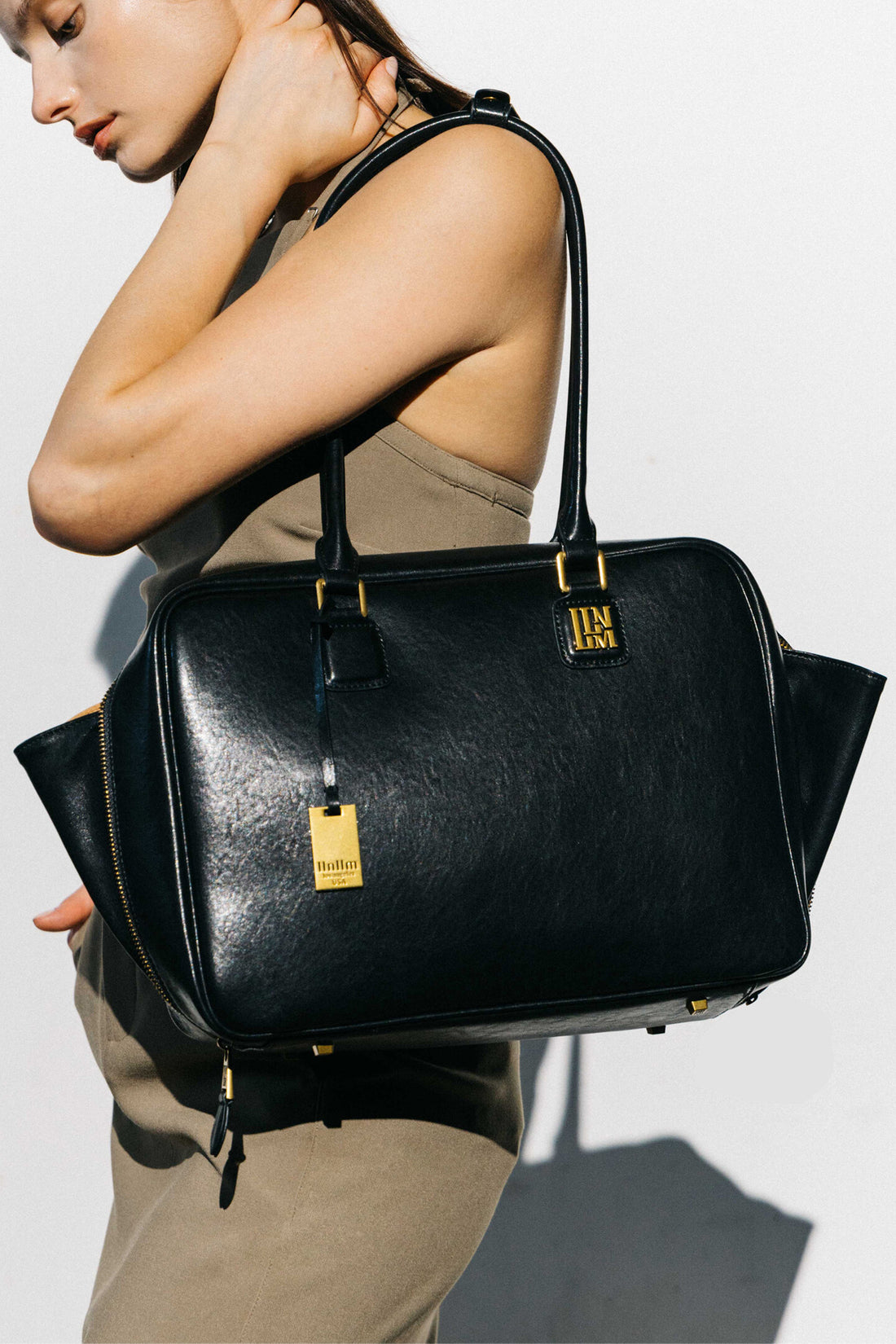 Model carries llnllm black leather satchel with structured double top handles, gold logo hardware, and side zipper details for a polished everyday look.