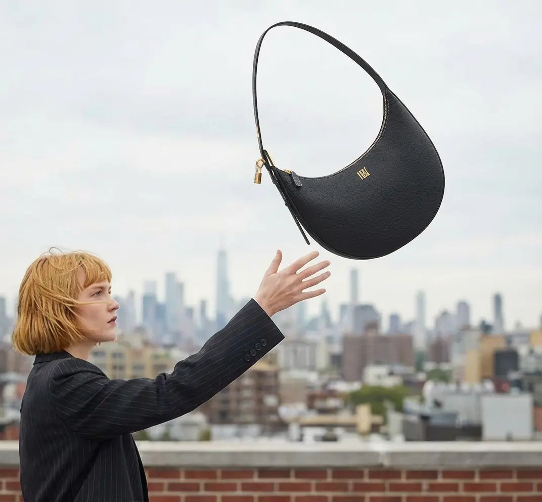 Black minimalist leather shoulder bag floating above city skyline, crescent shape luxury handbag with gold hardware in modern architectural setting